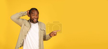 Foto de Cheerful surprised middle aged african american male in casual show finger to copy space, recommend sale, isolated on yellow background, panorama, studio. Advice, ad and offer, huge sale and idea - Imagen libre de derechos