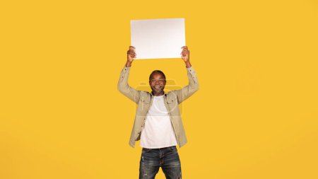 Téléchargez les photos : Happy handsome middle aged african american man in casual hold banner above his head with empty space isolated on yellow background, studio, panorama. Recommendation, advice, huge sale, ad and offer - en image libre de droit