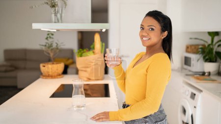Téléchargez les photos : Smiling millennial african american lady in casual holding glass of water in white modern kitchen interior with bag of vegetables, panorama. Weight loss and diet, aqua bananas and health care at home - en image libre de droit