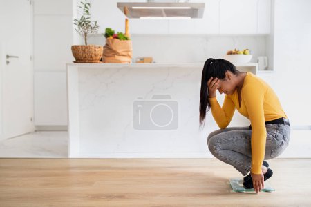 Téléchargez les photos : Sad millennial african american woman in casual crying on scales, suffering from excess weight in white kitchen interior. Slimming and health problems, stress and depression, weight loss at home - en image libre de droit