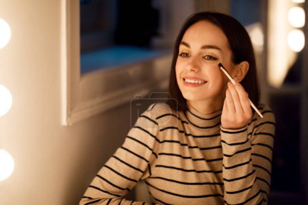 Téléchargez les photos : Happy cheerful beautiful brunette young lady in casual sitting in front of mirror, aplying makeup at home, holding visage brush and using eyeshadows or highlighter, smiling, copy space - en image libre de droit