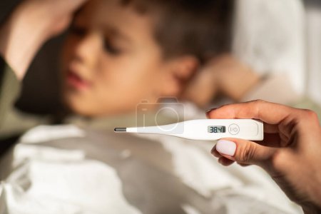 Photo for Tired little european boy lies on bed, sleeping, mom measures temperature with thermometer in bedroom, close up, blurred. Health problems, mother care, flu and cold, fever at home, treatment diseases - Royalty Free Image