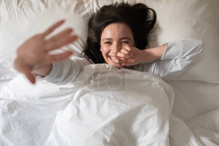 Foto de Cheerful pretty young european woman wakes up, waves her hand or closes from camera, lies on white bed in bedroom, top view. Sleep, health care, good morning at home, enjoy weekend and free time - Imagen libre de derechos