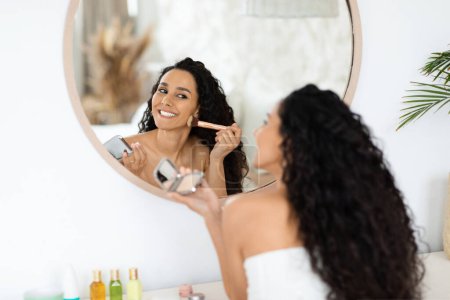 Téléchargez les photos : Smiling pretty arab millennial curly lady in towel applying blush on face, looking in mirror, enjoy daily procedure in bedroom interior. Good morning at home alone, beauty care and nude basic makeup - en image libre de droit
