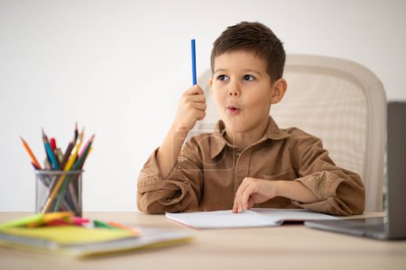 Photo for Cheerful cute little boy sits at table, studies, looks at pencil and got idea, enjoy learning and art in room interior. Lesson, education and childhood, creative and school, homework at home alone - Royalty Free Image