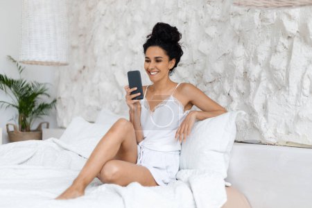 Téléchargez les photos : Smiling middle eastern millennial curly lady in domestic clothes woke up, sit on white bed, typing on smartphone in bedroom interior. Good morning alone at home with new app, blog and social networks - en image libre de droit