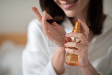 Téléchargez les photos : Cheerful young european woman on white bed applies spray on hair in bedroom interior, close up, cropped, blurred. Beauty care, treatment of dry hair and split ends at home, cosmetics for procedures - en image libre de droit