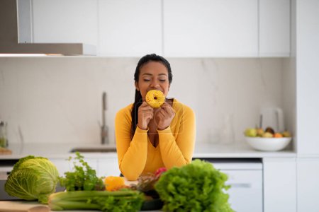 Foto de Cheerful hungry millennial african american lady in casual hold donut, enjoy aroma at table with organic vegetables in white kitchen interior. Weight loss, proper nutrition, health care or sweets - Imagen libre de derechos