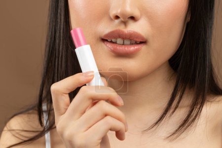 Photo for Cropped image of young asian lady holding pink lipstick in hand near face, japanese woman with nude lips standing over brown background, studio shot, closeup - Royalty Free Image