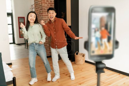 Téléchargez les photos : Excited asian lifestyle bloggers filming video on cellphone, dancing on phone camera at home. Happy spouses live streaming for entertainment blog, shooting for social network - en image libre de droit