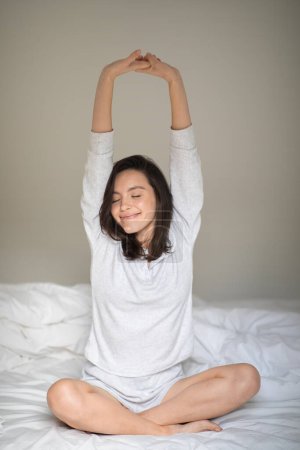 Téléchargez les photos : Happy pretty young european woman in pajamas woke up, stretching body, enjoys free time, sitting on white bed in bedroom, vertical. Sleep, health care, rest and relax at home, weekend and good morning - en image libre de droit