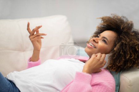 Photo for Happy cheerful smiling beautiful young african american woman in comfy casual outwear lying on couch, gesturing while having phone conversation with friend or lover at home, copy space - Royalty Free Image