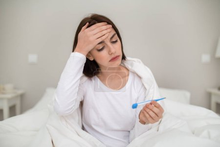 Photo for Unhappy sick young brunette woman sitting in bed covered with warm duvet, touching forehead and holding electronic thermometer, suffering from fever while having cold or flu, copy space - Royalty Free Image