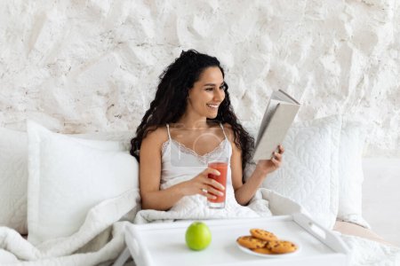 Photo for Cheerful arabic millennial curly lady in domestic clothes woke up, sit on bed, has breakfast and reads book in bedroom interior. Good morning alone at home, female enjoys hobby in free time with food - Royalty Free Image
