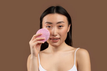 Foto de Face cleansing concept. Attractive young japanese woman holding soft pink cellulose sponge, washing her face, posing isolated on brown background, studio shot - Imagen libre de derechos