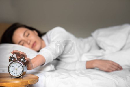 Foto de Tired sad unhappy young european woman in pajamas wake up, turns off alarm clock, lies on white bed in bedroom. Problems with sleep, health care, time for work and meeting, early morning at home - Imagen libre de derechos