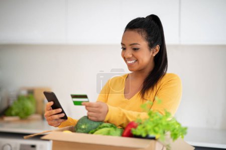 Foto de Glad millennial african american woman unpacks cardboard box with organic vegetables uses smartphone and credit card in white kitchen interior. Ordering grocery and delivery, household chores at home - Imagen libre de derechos