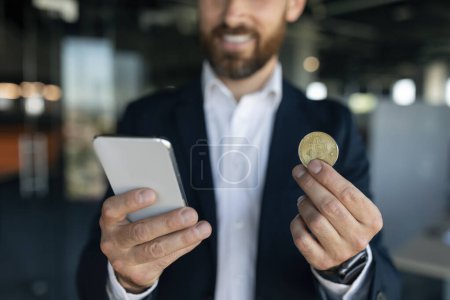 Téléchargez les photos : Online trading. Happy middle aged businessman holding golden bitcoin and mobile phone, man earning cryptocurrency, standing in office interior, selective focus - en image libre de droit