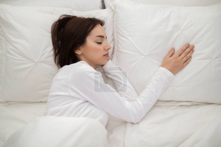 Foto de Loneliness concept. Young brunette woman lying in double bed with one hand on empty pillow next to her, miss her boyfriend, lady wearing white cotton pajamas, top view, copy space - Imagen libre de derechos