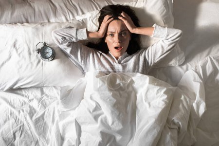 Photo for Shocked young european woman in pajamas woke up, holding her head, lies on white bed with alarm clock in bedroom, top view. Problems with sleep, health care, overslept for work and meeting, stress - Royalty Free Image