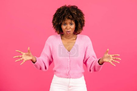 Téléchargez les photos : Shocked confused millennial african american curly female in casual with open mouth spreads her arms to sides isolated on pink background, studio. Amazing bad news, ad and offer, emotions, problems - en image libre de droit