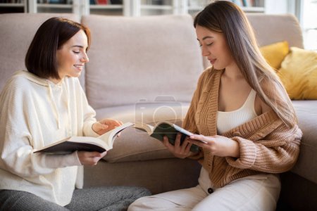 Photo for Cheerful pretty european millennial ladies in sweaters read books, talk, enjoy rest, relax and free time in comfort living room interior. Study, knowledge and education together, family relationships - Royalty Free Image
