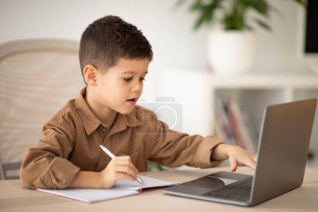 Téléchargez les photos : Serious busy little boy sits at table, studies, has video call on laptop, makes notes in room interior. Homework, remote study lesson, upbringing and childhood, new normal at school during covid-19 - en image libre de droit