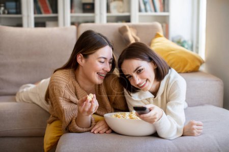 Foto de Cheerful millennial european girlfriends in sweaters lies on sofa with remote control, enjoy popcorn in free time, watch tv in comfort living room interior. Fun together, movie night at home and sneck - Imagen libre de derechos