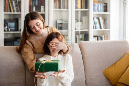Foto de Cheerful european millennial lady closes eyes to her friend and gives gift box in living room interior, copy space. Celebration holiday, anniversary, birthday surprise and congratulations at home - Imagen libre de derechos