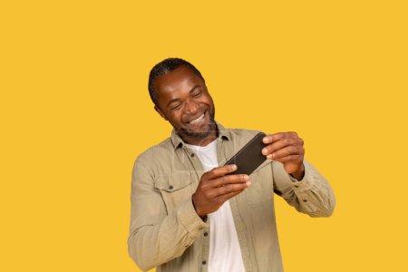 Téléchargez les photos : Happy middle aged african american male in casual play online game on phone, enjoy spare time, isolated on yellow background, studio. App for entertainment and fun with device, winning, ad and offer - en image libre de droit