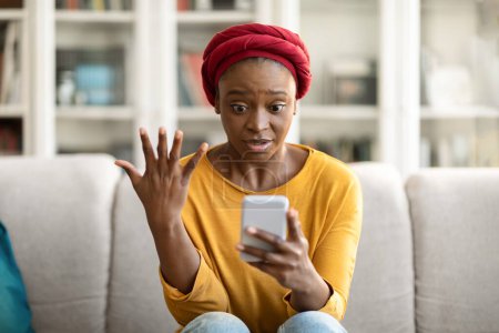 Téléchargez les photos : Stressed anxious young black woman in casual outfit wearing red turban sitting on couch in living room, holding modern smartphone and gesturing, home interior, checking her bank account, copy space - en image libre de droit
