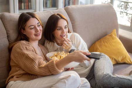 Photo for Glad european millennial ladies in sweaters sit on sofa with remote control, enjoy popcorn and watch film on tv in free time in comfort living room interior. Relationship, movie night together at home - Royalty Free Image
