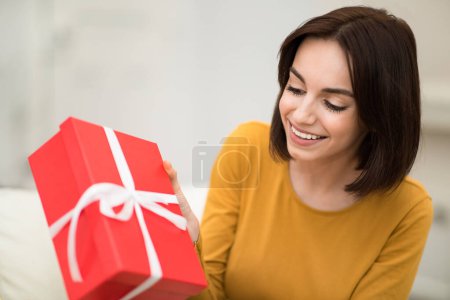 Téléchargez les photos : Cheerful pretty brunette young woman sit on couch in living room unpack red cardboard box birthday present, smiling excited millennial lady open gifts after winter holidays celebration, copy space - en image libre de droit
