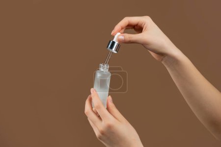 Photo for Closeup shot of young lady holding moisturizing serum, standing on brown background. Unrecognizable woman nourishing her skin, using hyaluronic acid, pampering herself at home - Royalty Free Image