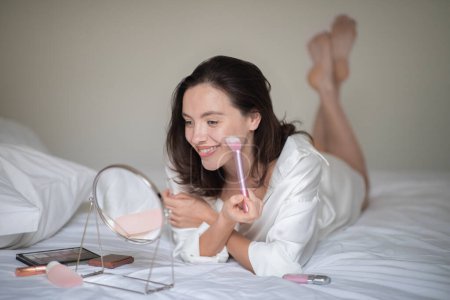 Foto de Cheerful young european lady in bathrobe lies on white bed, apply cosmetics with brush on face, looks in mirror in bedroom interior. Beauty care, natural nude makeup at home, lifestyle at spare time - Imagen libre de derechos
