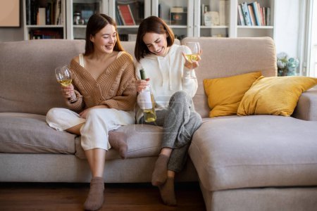 Photo for Laughing european millennial ladies in sweaters with glasses of wine, bottle enjoy rest, relax, party at free time in comfort living room interior. Evening together, date and holiday celebrate at home - Royalty Free Image