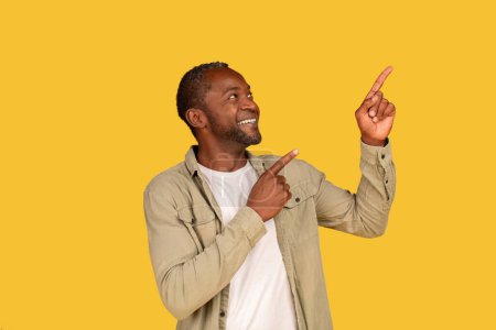 Téléchargez les photos : Recommendation, advice and look at here. Glad middle aged african american man in casual pointing his fingers at empty space, isolated on yellow background, studio. Sale, ad and offer, great news - en image libre de droit