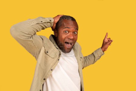 Téléchargez les photos : Portrait of satisfied surprised mature black man with open mouth, hold hand to head, show finger to free place, recommend huge sale, isolated on yellow background. Emotions of victory and success - en image libre de droit