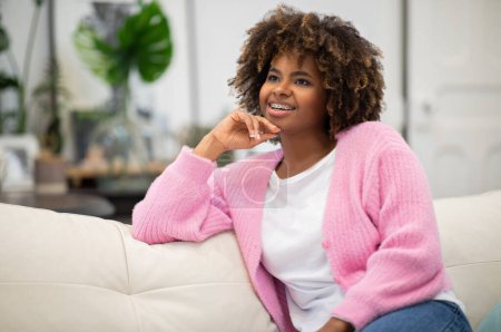 Foto de Dreamy cheerful stylish african american attractive curly millennial woman in comfy outwear resting on couch in living room, looking at copy space for ad and smiling, spending weekend at home - Imagen libre de derechos