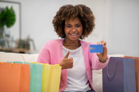 Téléchargez les photos : Unlimited shopping, online sales. Overjoyed emotional pretty young african woman customer sit among shopping bags at home satisfied with great purchase, showing credit card and thumb up, copy space - en image libre de droit