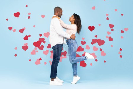 Téléchargez les photos : Love and care. Happy black couple hugging and looking at each other, standing on blue studio background with red flying hearts, full length shot, side view. St. Valentines Day - en image libre de droit