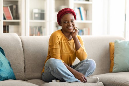 Foto de Cheerful happy relaxed young african american woman in casual with red headscarf on her head chilling alone at home, sitting on couch with legs up, smiling at camera, cozy living room interior - Imagen libre de derechos