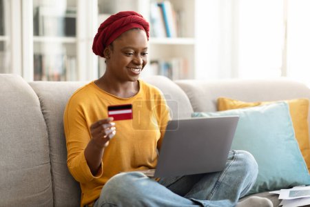 Téléchargez les photos : Happy attractive millennial african american woman shopping from home, sitting on couch in cozy living room, using laptop and credit card, copy space. Online sales, e-commerce, retail concept - en image libre de droit