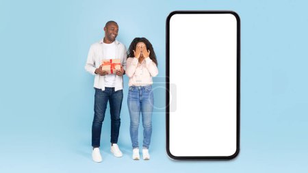 Photo for Excited african american couple near big smartphone with empty white screen, man holding gift box, happy lady covering her eyes with hands and waiting for surprise, mock up collage - Royalty Free Image