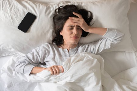 Téléchargez les photos : Sad young european woman wakes up, presses her hand to head, suffers from headache, has bad dreams, lies in white bed in bedroom, top view. Problems with sleep, health care, early morning at home - en image libre de droit