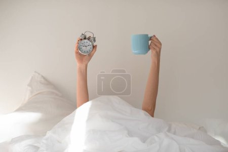 Foto de Hands of young european woman under covers with alarm clock, cup of coffee on white bed in bedroom, free space, unrecognizable. Good morning, energy, sleep well, health care, time for work, meeting - Imagen libre de derechos