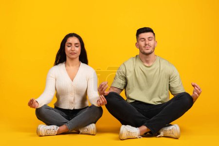 Foto de Cheerful relaxed young arabic woman and guy in casual with closed eyes sit in lotus position meditating, enjoy silence and peace, isolated on yellow background, studio. Rest, break, ad and offer - Imagen libre de derechos