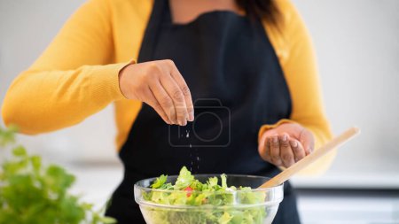 Photo for Millennial african american woman in apron salting salad, cooking dinner at table with organic fresh vegetables in white kitchen interior, close up, cropped. Household chores, veganism, health care - Royalty Free Image