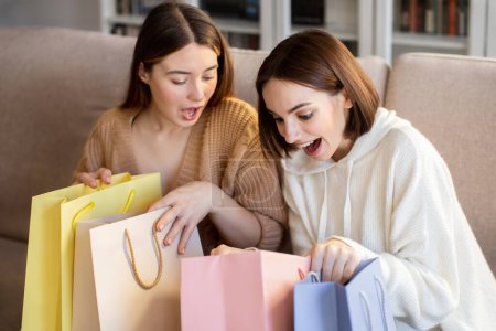 Téléchargez les photos : Happy surprised european millennial girlfriends shopaholic in sweaters open bags from store, enjoy free time, sale and purchases in comfort living room interior. Reaction on buy, shopping at home - en image libre de droit