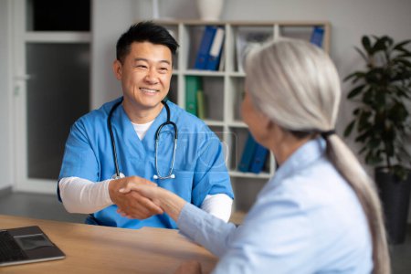 Téléchargez les photos : Cheerful middle aged korean male doctor shaking hands with old caucasian woman patient in clinic office interior. Greeting, hello, health care, treatment disease and visit to therapist in hospital - en image libre de droit